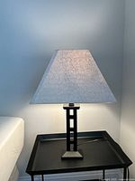 Black metal table lamp with square tan fabric shade placed on a black tray table, photographed from a slight angle showing on position and lit shade.