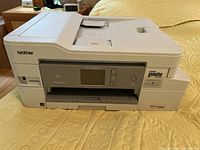 Front and side view of the white Brother Work Smart Series MFC-J995DW printer on a yellow textured bedspread, showing buttons, display and paper feeds.