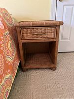 Front view of coastal wicker side table featuring one drawer and open shelf below, natural brown color, placed next to a colorful bedspread on carpet.