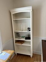 Front view of tall cream wooden bookshelf standing against wall with some office and household items on shelves.