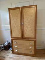 Front view of light brown wooden armoire with decorative inlaid door panels and three drawers beneath, showing white handles.