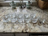 Top view of the 11 clear glass tumblers with etched Harvard and Cambridge scenes aligned on a granite countertop.