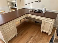 Front and left angled view of L-shaped farmhouse desk showing cream panel base and brown wood top with drawers and cabinet doors.
