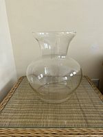 Front view showing rounded body and flared lip of large clear glass vase placed on wicker surface.