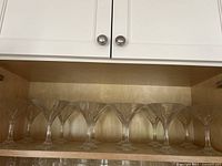Wide view of 12 clear patterned glass martini glasses arranged on a wooden shelf inside a cabinet.