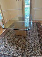 Angled view of rectangular clear glass tabletop with polished edges on wooden pedestal base, placed on patterned rug in bright room.