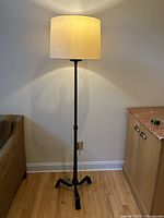 Single black metal floor lamp with beige drum shade, turned on and plugged in near sofa and cabinet.