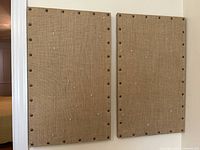 Two tan fabric-covered bulletin boards side by side, showing studded edges and pin holes on light-colored wall.