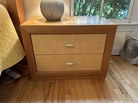 Front view of one wood night stand showing 2 drawers with light wood inlaid panels and silver oval handles, sitting on a hardwood floor near a window.