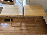 Two light wood folding tables shown side by side on a wood floor, tabletop surfaces visible with slight wear and scratches.