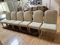 Six cream fabric and wood dining chairs arranged in a row on a hardwood floor next to a patterned rug in living room.