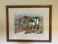Frontal view of the signed watercolor landscape painting in gold wood frame with matting, showing houses, flowers, greenery, and background mountains.