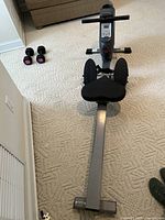 Front view showing the footrests, seat, handlebar, and LCD display of the rowing machine with dumbbells on the floor in background.