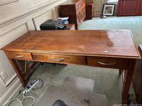 Front and side view of brown wooden office desk with three drawers and electrical sockets, showing surface wear and scratches.