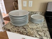 Photo showing stacked white dinner plates and salad plates with black rims on a kitchen counter.