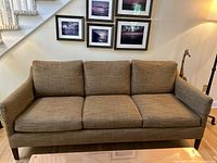 Front view of brown fabric sofa with three cushions, framed pictures on wall behind the sofa.