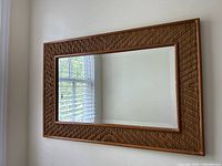 Photo showing the entire rectangular wall mirror with the woven design brown wooden frame mounted on a white wall, reflecting window blinds.