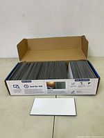 Photo of open box containing 102 glossy white peel and stick subway wall tiles measuring about 3x6 inches, with one tile shown outside the box.