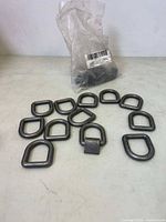 Plastic bag containing multiple black forged steel D rings with attached welding clips, packaging partially open.