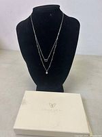 Double layered silver necklace displayed on black bust stand next to white box with butterfly logo and text.