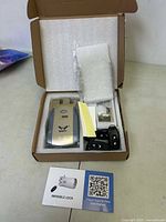Open box showing main lock unit, remote controls, mounting hardware, and instruction card inside foam-lined packaging.