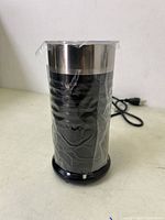 Front diagonal view of the black and stainless steel Keurig milk frother, still wrapped in plastic and plugged in with black power cord visible.