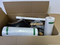 View inside open cardboard box showing multiple rolls of vacuum seal bags, a speckled frying pan, and other kitchen items.