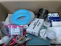 Top-down view inside box showing blue kitchen container, metal mug, pink rope, and various small packaged items including toolsure box and small metal tin.