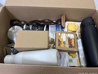Open cardboard box showing assorted kitchen and household items including thermos bottles, silicone molds, metal strainer, and food saver boxes.