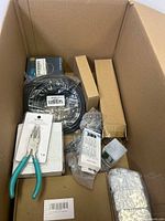 Large cardboard box containing various new miscellaneous tools, electronics cables, and accessories including pliers, coveralls, and small sealed parts.