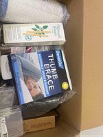 Top view of a box with packaged items including GeneGo thumb brace, Miswak brush box, pink compact, and decorative bottle.