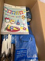 Box opened showing colorful party decor pad with 'Cheers' banner, cones, cupcake toppers, white plastic forks, brown paper cups, and blue plastic table cover.