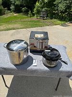 Photo showing the 16-quart stainless steel stock pot with lid and the separate electric pressure cooker with lid and power cord on a gray surface outside.