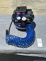 Full view of the air compressor showing the blue tank, black motor section, attached blue coiled hose with brass fittings, and air blow gun attachment.