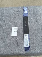 Single 22-inch mower blade in black metal, sealed in blue and white branded packaging with product label visible, placed on grey fabric surface next to auction number card.