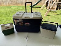 Photo of three coolers on a table outside: a large Coleman cooler with beige lid and two smaller coolers on either side.