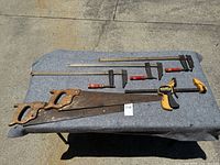 4 clamps and 2 hand saws laid out on a gray cloth, displayed on concrete ground. Clamps include metal bar clamps with different colored handles, and hand saws have wooden handles with visible wear.