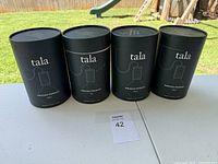 Photo of 4 black cylindrical boxes labeled 'tala Knuckle Pendant' on a table outdoors.