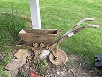 Side view of antique garden seeder showing wooden hopper, two wooden handles, metal rear wheel, and metal soil tilling blades attached underneath.