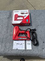 Red Arrow electric staple gun and nailer with power cord, original box, and instruction manual displayed on concrete surface.