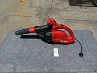 Red and black Black and Decker corded electric leaf blower laid on grey fabric outdoors, showing overall condition and attached power cord.