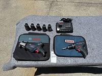Lot items displayed on outdoor grey cloth: five batteries, two Bosch branded cases, Bosch charger, Bosch cordless drill, and Bosch reciprocating saw.