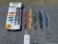 12 piece Cuisinart Advantage cutting utensil set displayed on grey surface. One knife remains in original packaging, others still in plastic blade covers with varied colored handles.