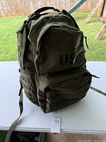 Front view of the olive green military-style backpack with 'US' marking on the front compartment and visible MOLLE webbing.