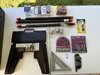 Top view of various tools displayed on a white table including metal pipes, foldable sawhorse, metal clamps, corner clamp, and bit sets.