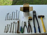 Full lot of tools on white table showing all items including wrenches, pliers, hammer, tape measure, and box of nails