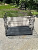 Full view of the large black metal wire dog crate with plastic tray on pavement outdoors with grass and bench in background.