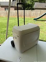 Full view of the large white rolling cooler with extended handle and wheels on a table outdoors.