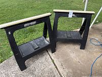 Pair of black plastic sawhorses with white wooden tops standing on pavement near grass.