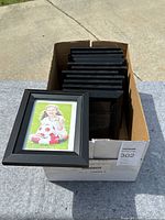 View of all 12 black frames stacked in a cardboard box outdoors on concrete surface.